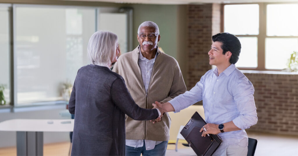 A diverse group in a bright office: a woman and a younger man smiling and shaking hands, while an older man watches, conveying friendliness and collaboration.