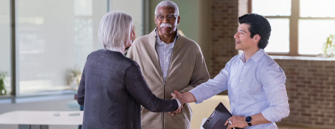 A diverse group in a bright office: a woman and a younger man smiling and shaking hands, while an older man watches, conveying friendliness and collaboration.